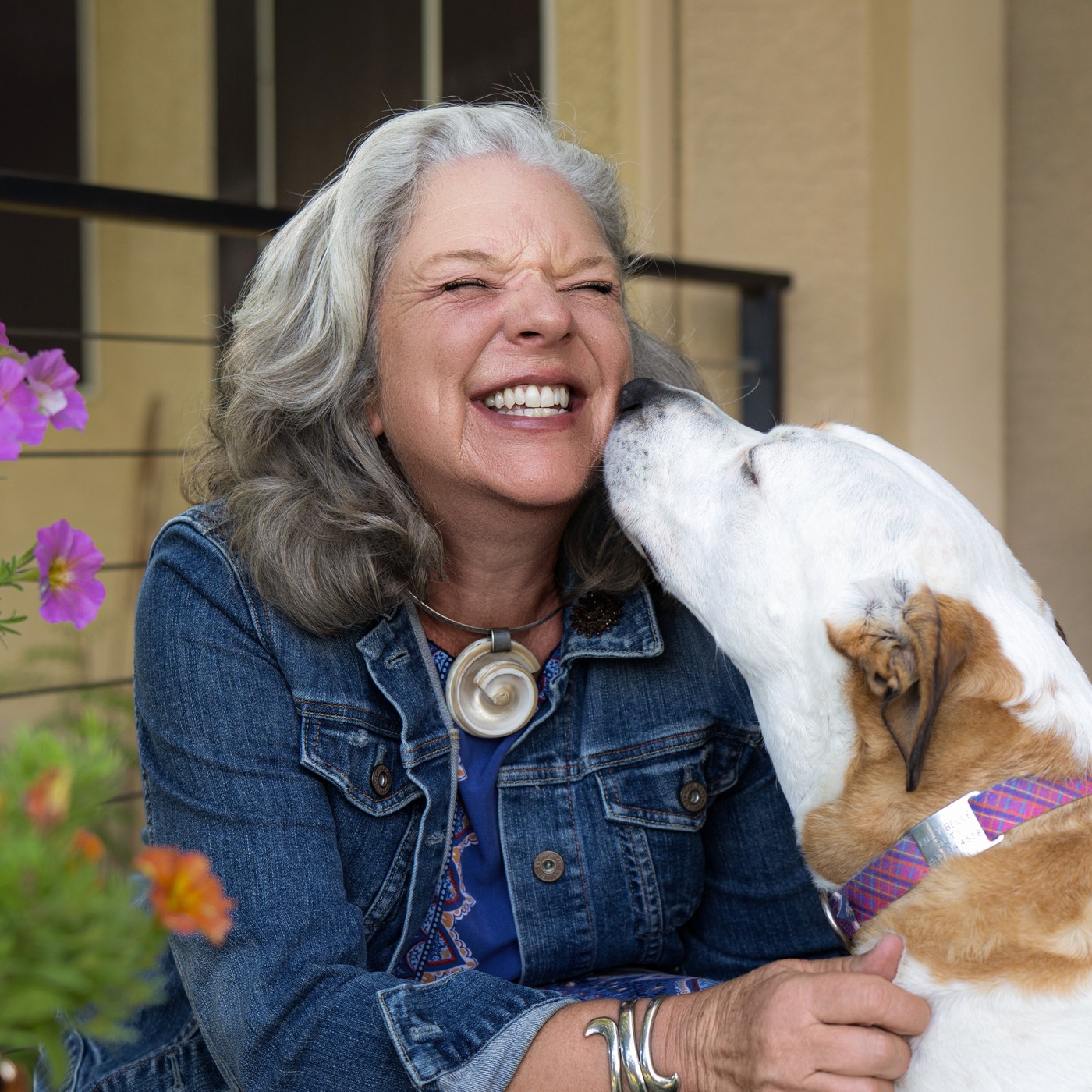 Laura laughing in a denim jacket while her dog nuzzles her cheek, petunias in the foreground