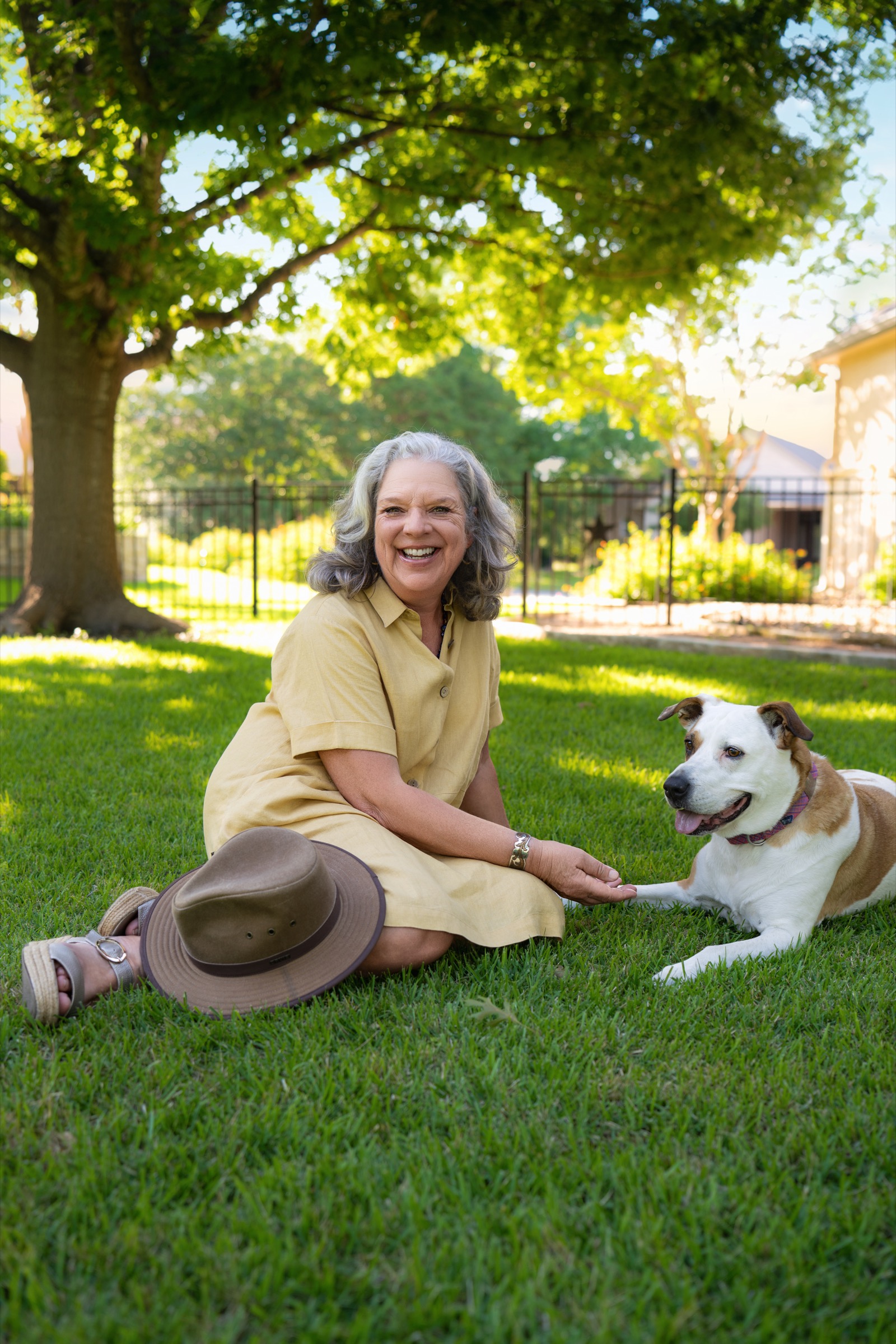 Laura sitting in the grass under a live oak at golden hour, laughing, her hand resting in her dog's paw